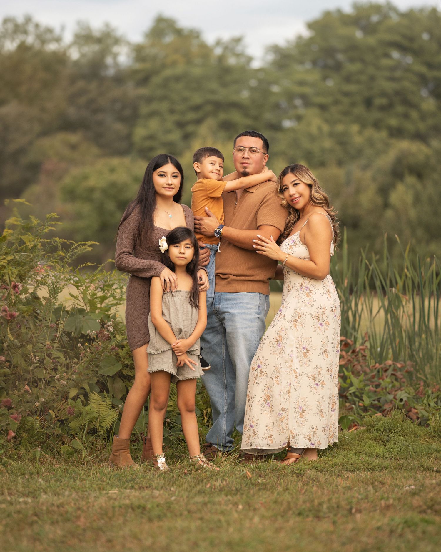 A group stands together outdoors for a family portrait against a natural backdrop of trees and tall grass at sunset.