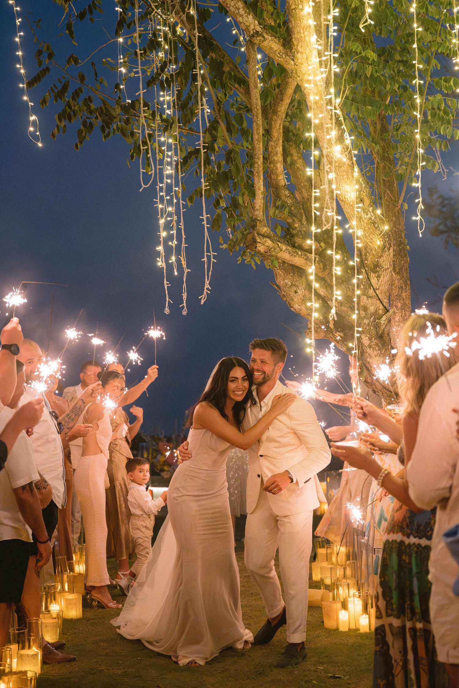 Wedding celebration at night with sparklers and string lights hanging from tree creating magical outdoor ambiance.