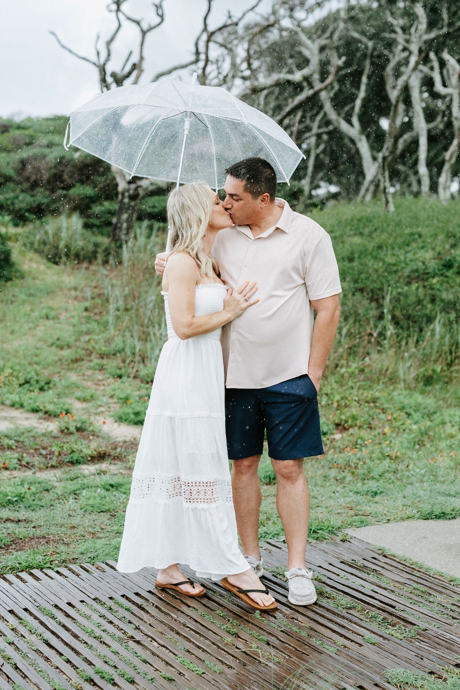 Romantic moment captured on a wooden boardwalk with clear umbrella and coastal scenery.