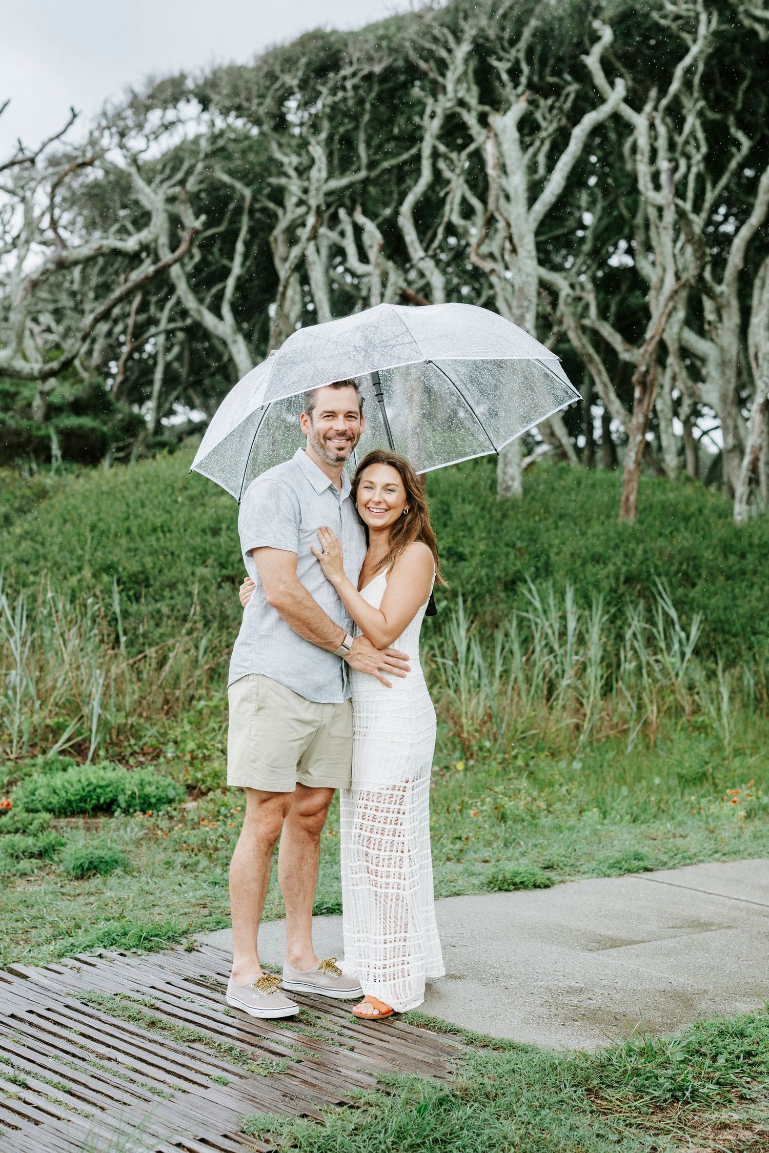 Couple sharing tender moments under a clear umbrella on a coastal walkway.