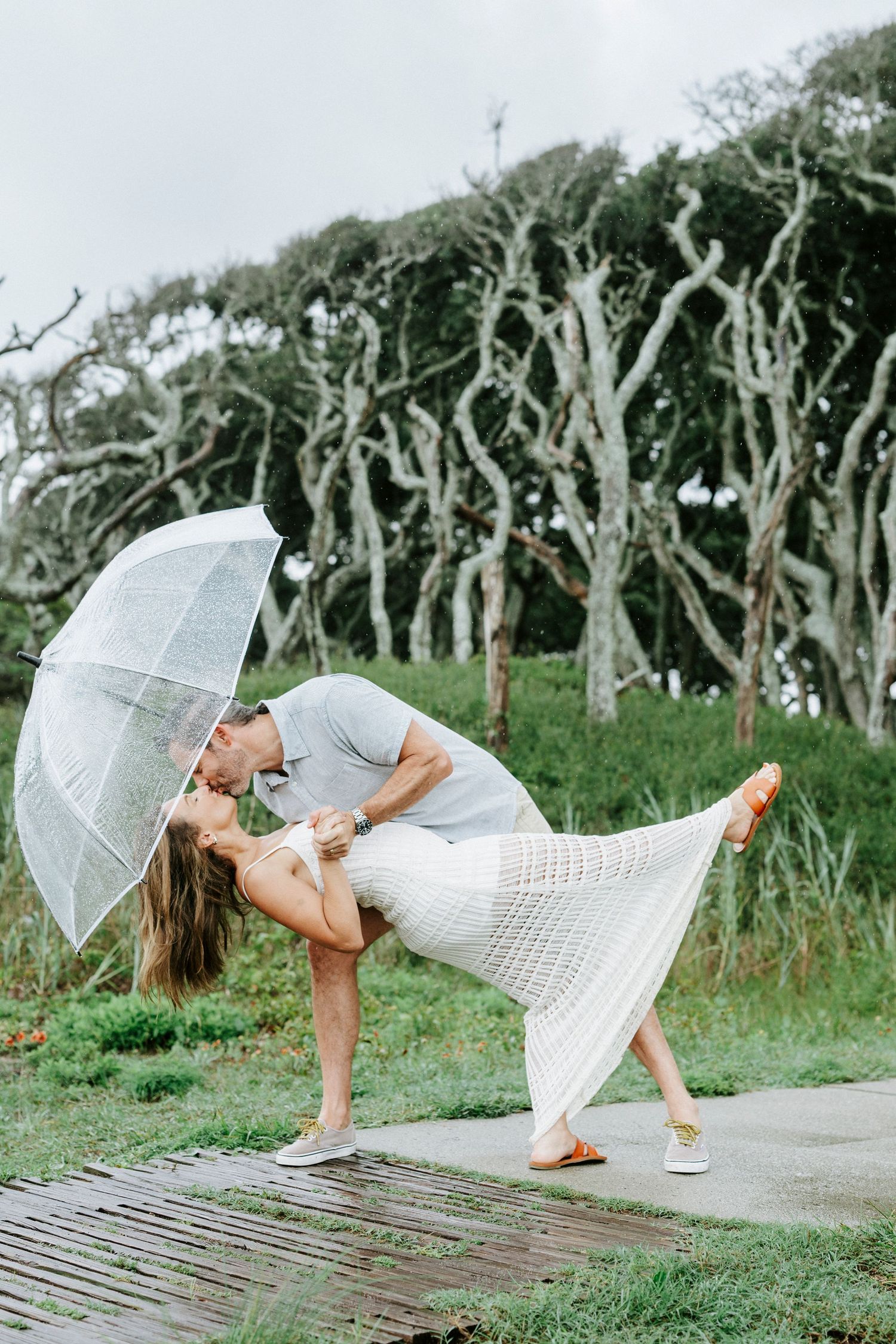 Playful dance poses with clear umbrella on a coastal pathway.