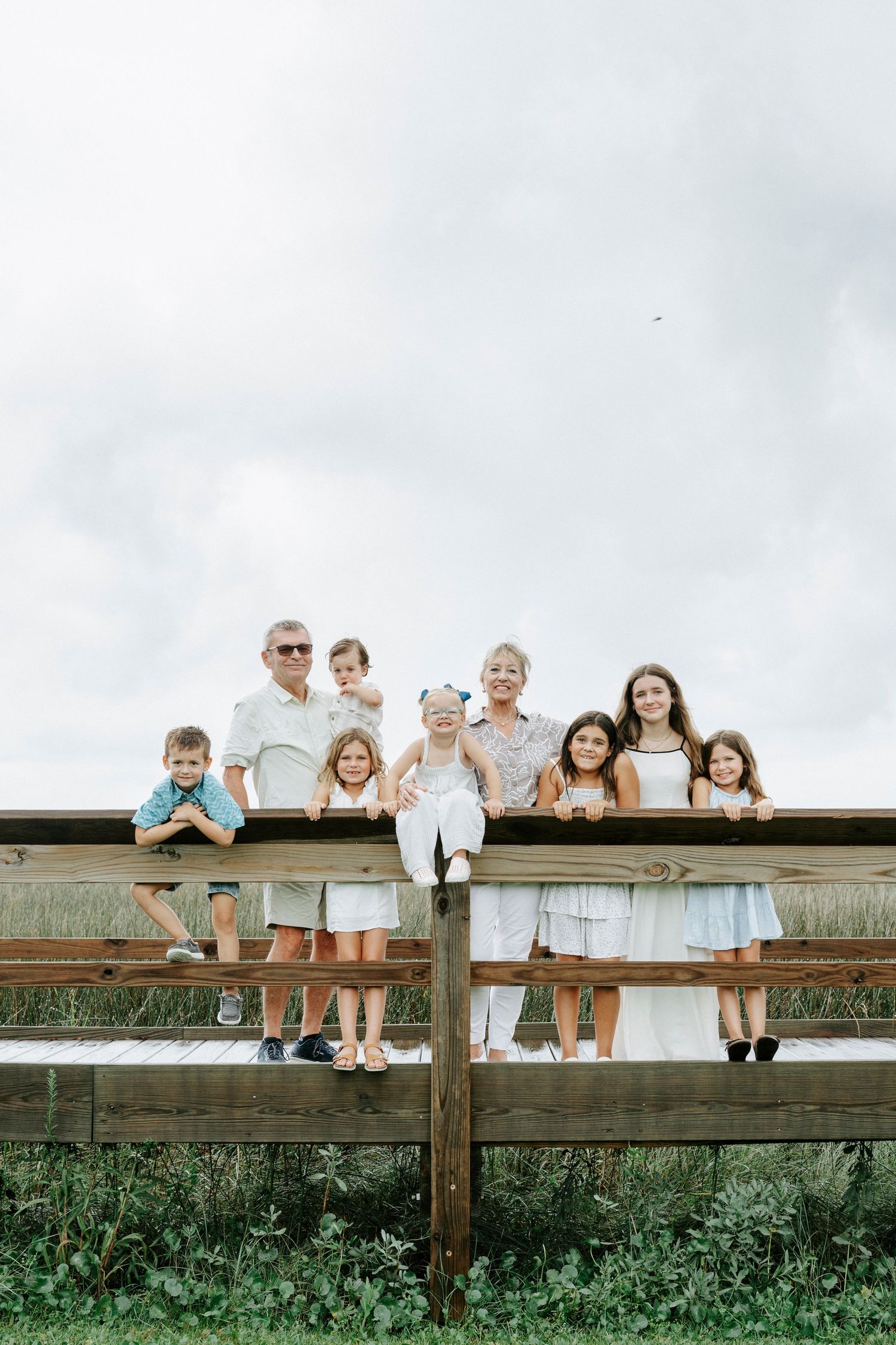 Large extended family sitting together on a wooden fence in casual summer attire against a cloudy sky background.