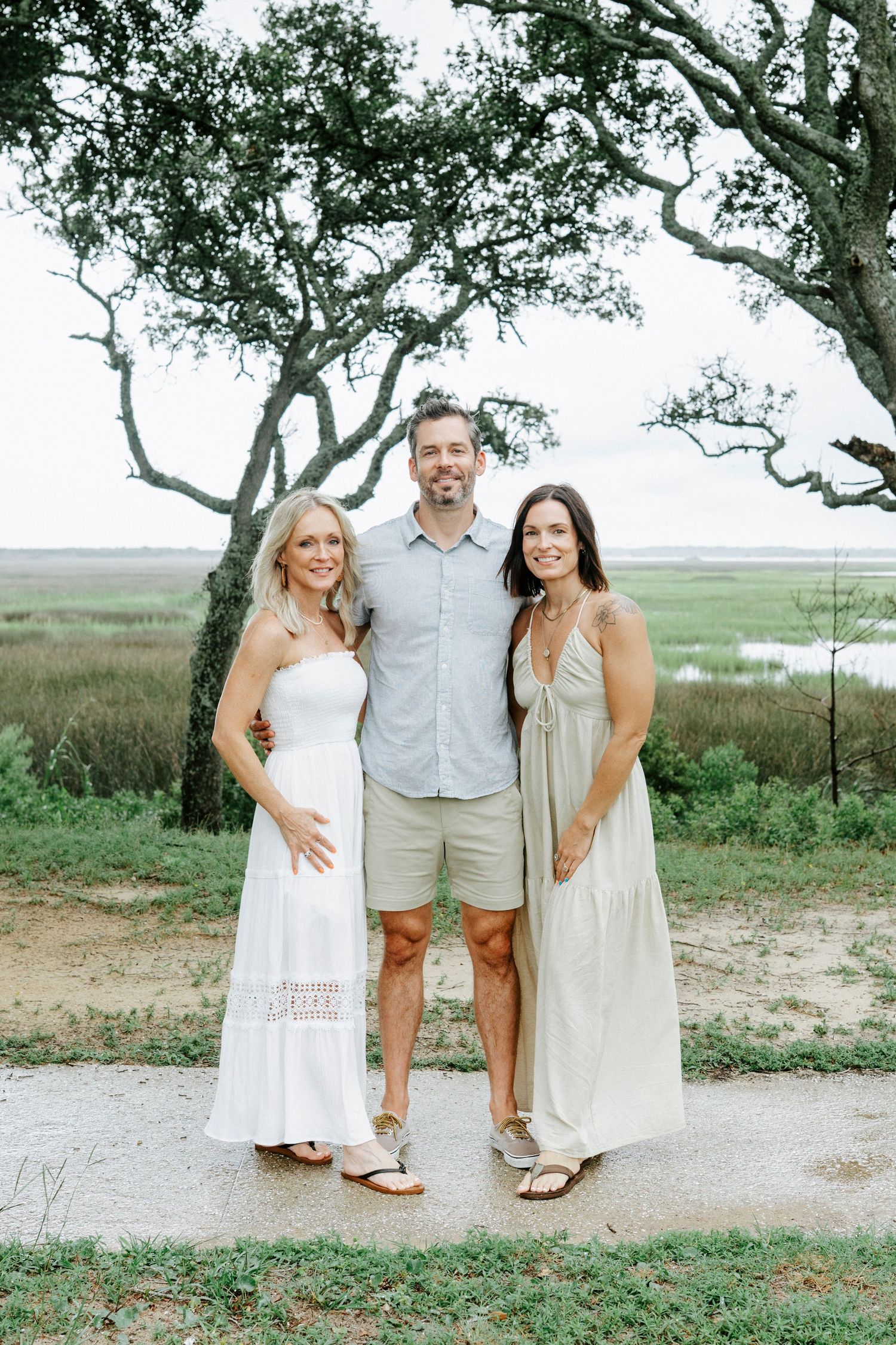 Three people in light summer clothing pose together under a tree with marshland in the background.