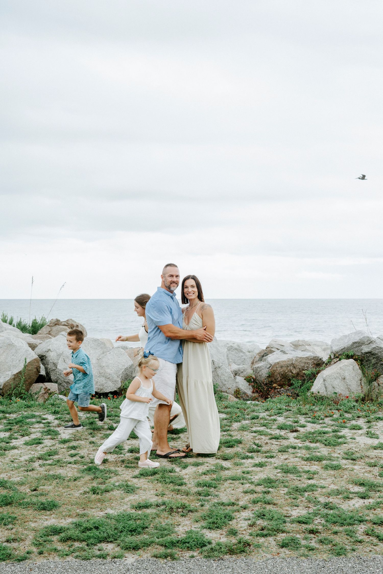 Family members play and interact on grassy area near rocky coastline.