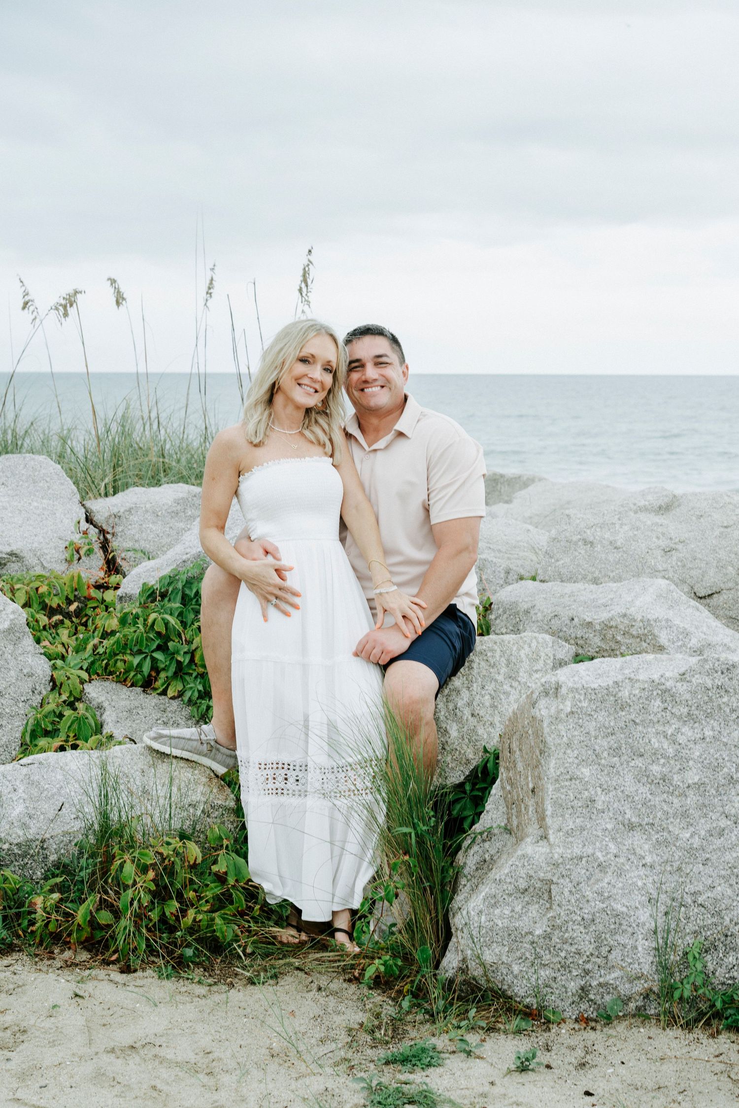 Couple sits together on large coastal rocks with ocean waves in background.