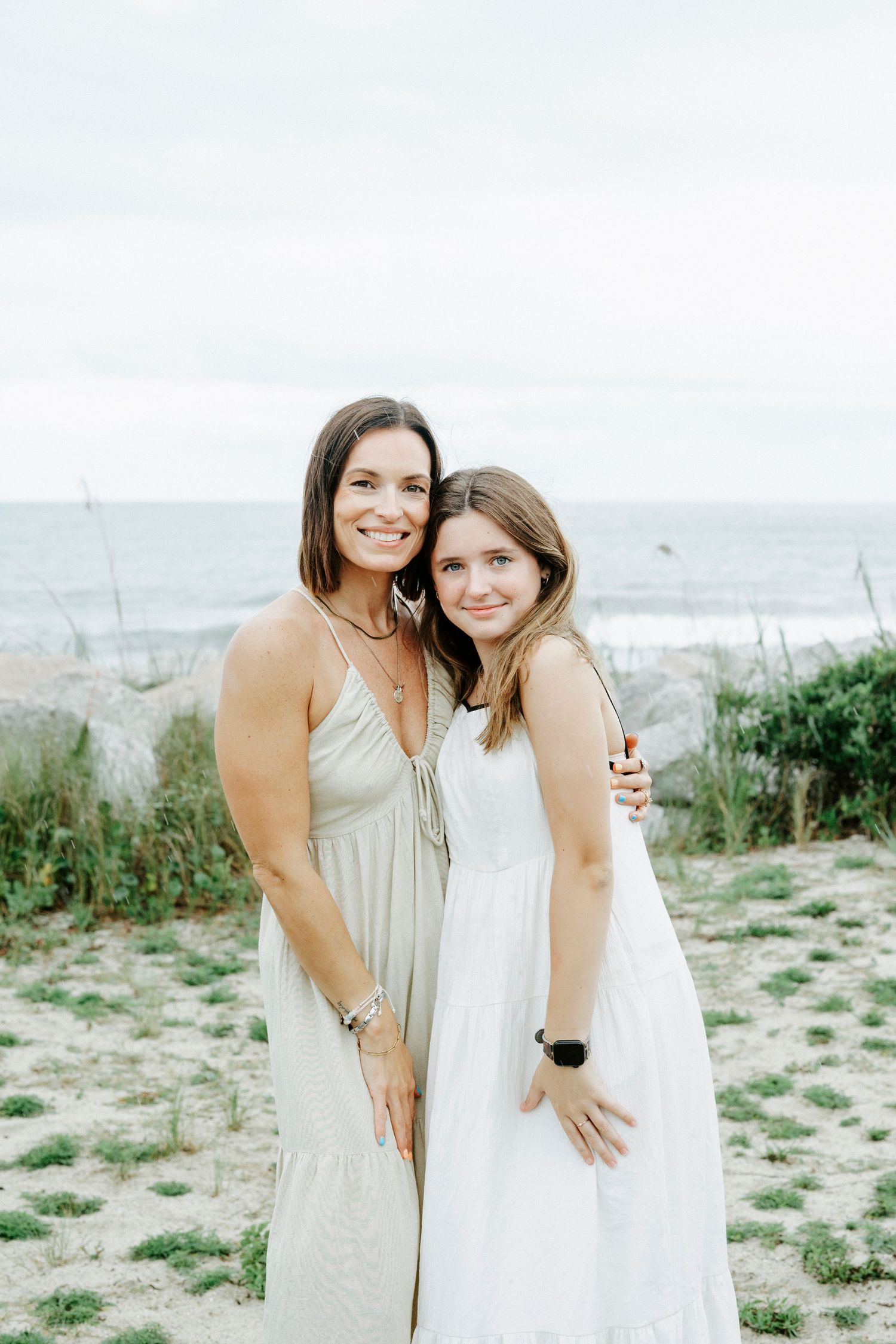 Two people in light colored dresses embrace on beach with ocean in background.