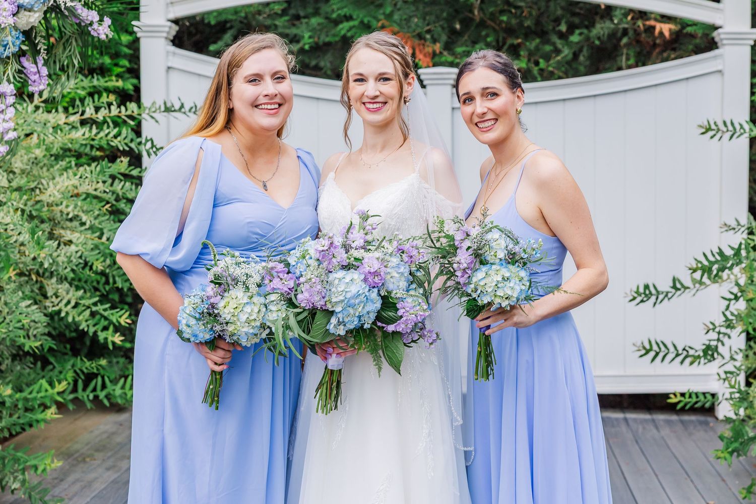 Wedding party members in matching periwinkle blue dresses hold purple and white floral bouquets at an outdoor celebration.