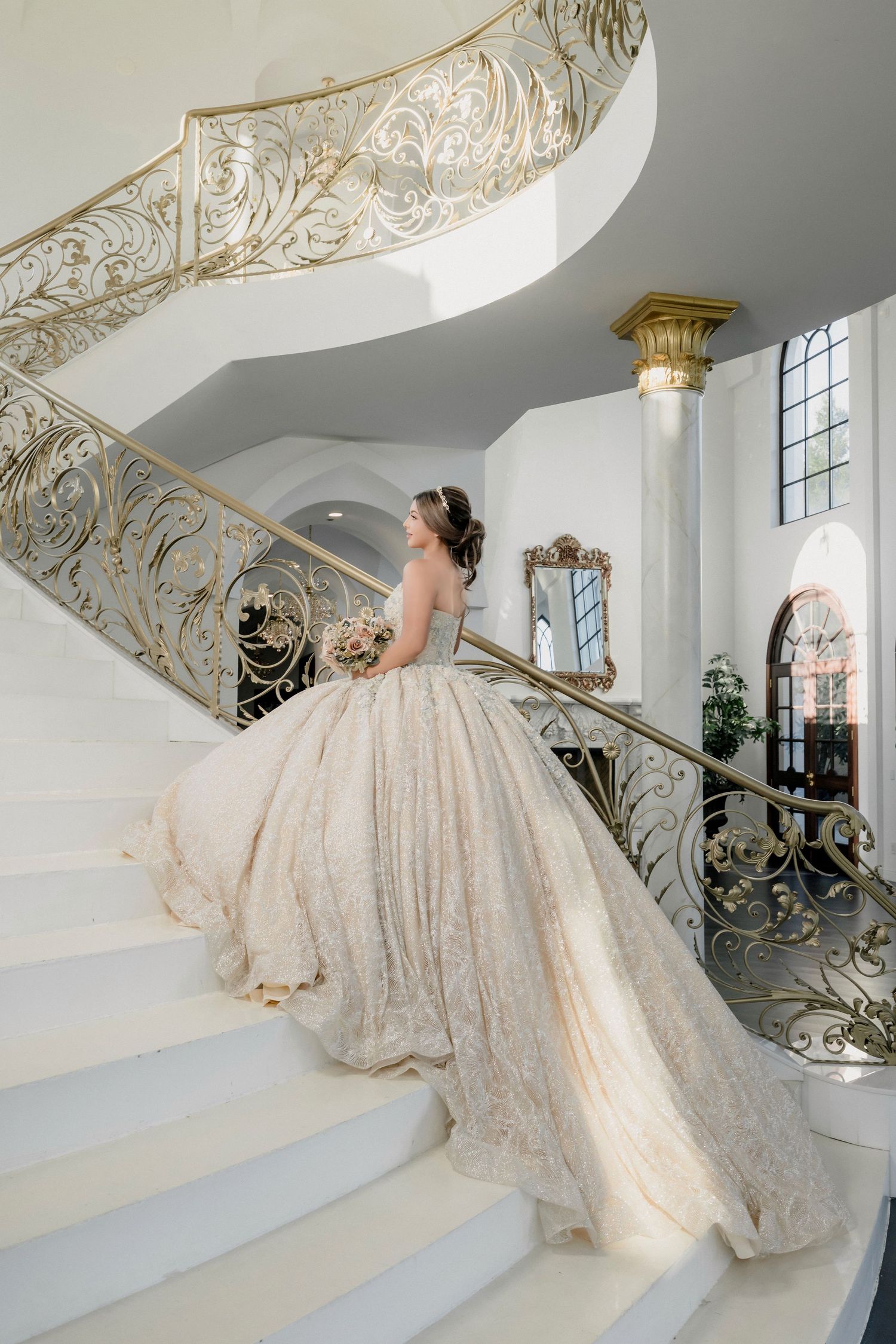 Elegant ballgown wedding dress flowing down ornate marble staircase with gold filigree railing in luxury venue.