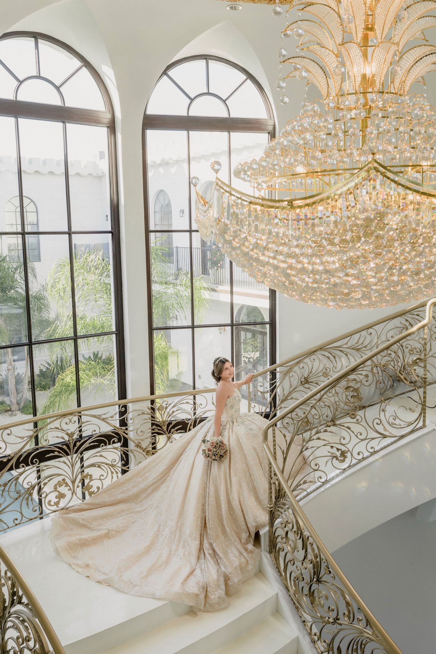 A luxurious wedding dress twirls in a grand room with crystal chandeliers and decorative gold stair railings.