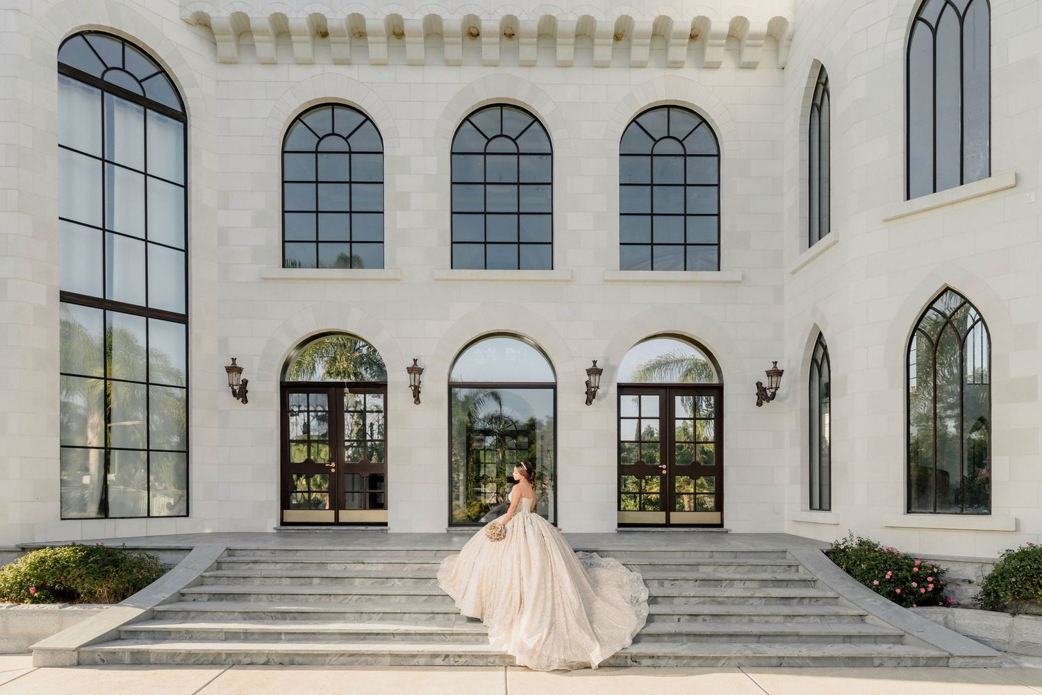 Flowing champagne-colored wedding gown captured against white architectural facade with arched windows.