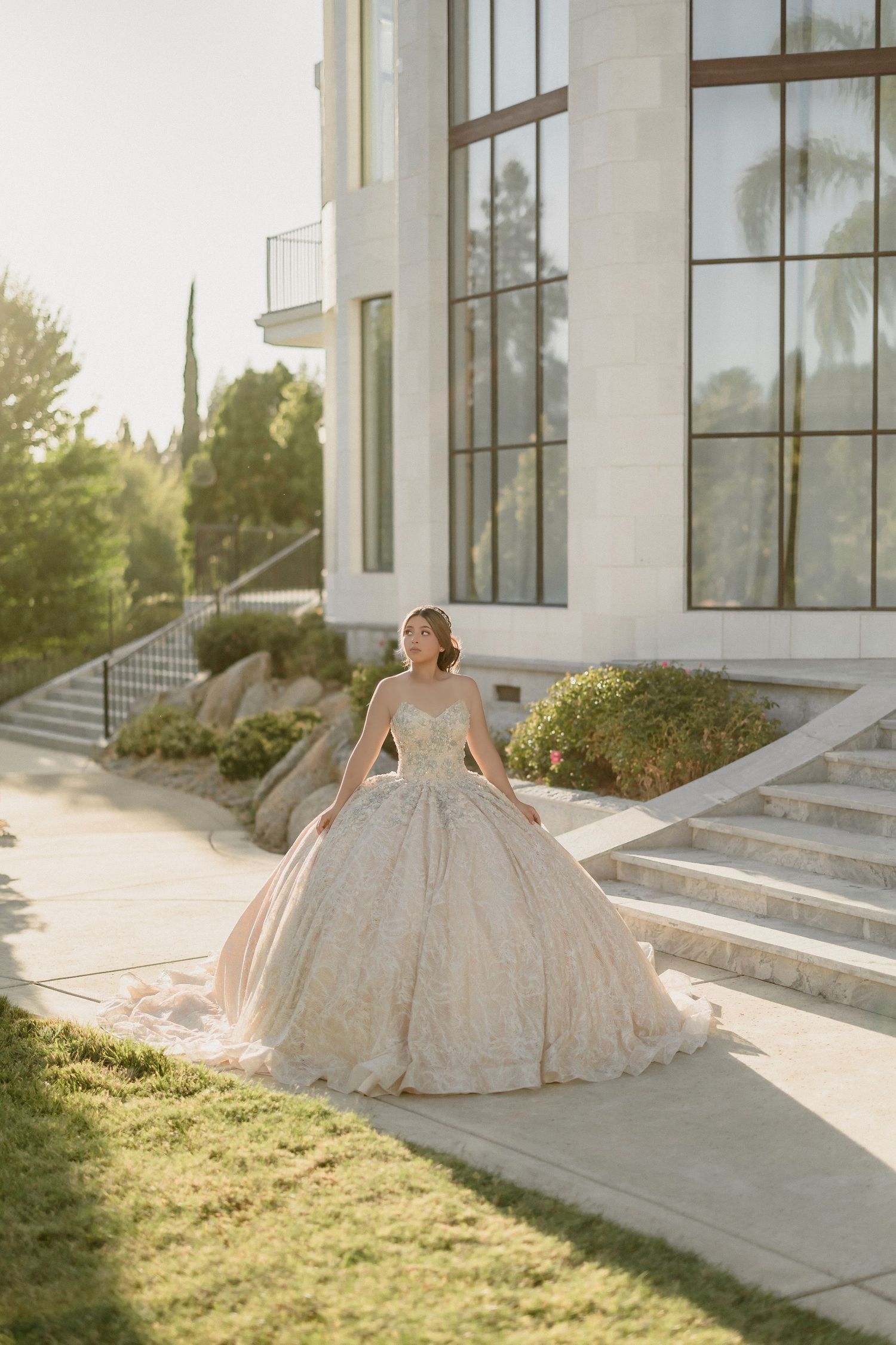 Elegant ballgown wedding dress glimmers in golden sunlight against modern white building architecture and green lawn.