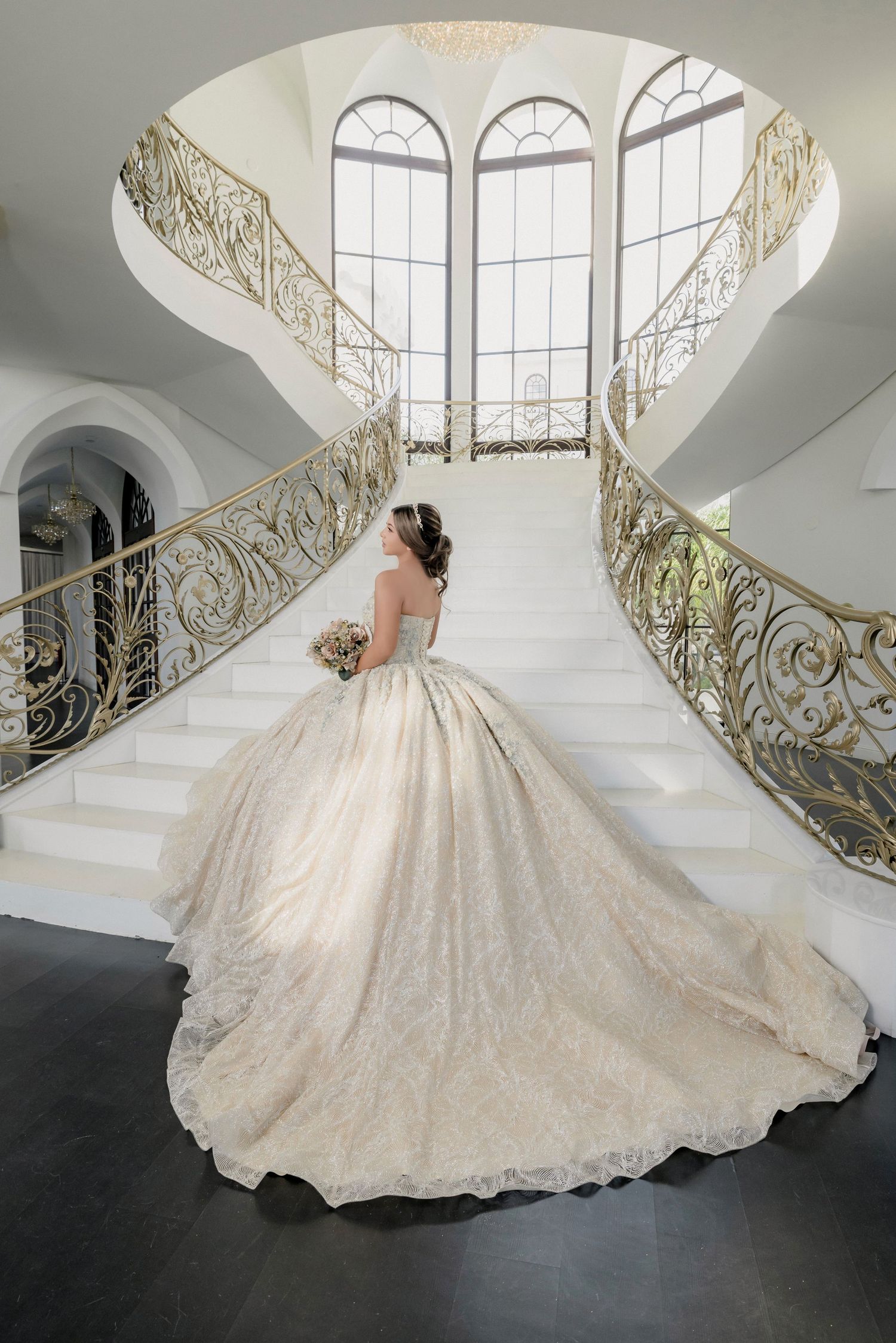 An ethereal bridal gown with a sweeping train glides across the floor near ornate golden railings and arched windows.