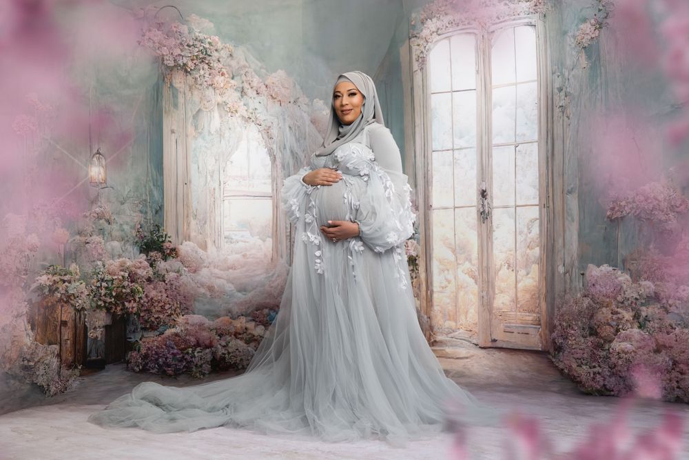 A pregnant woman in a flowing gray tulle gown poses in a dreamy floral setting with pink blossoms and French doors.