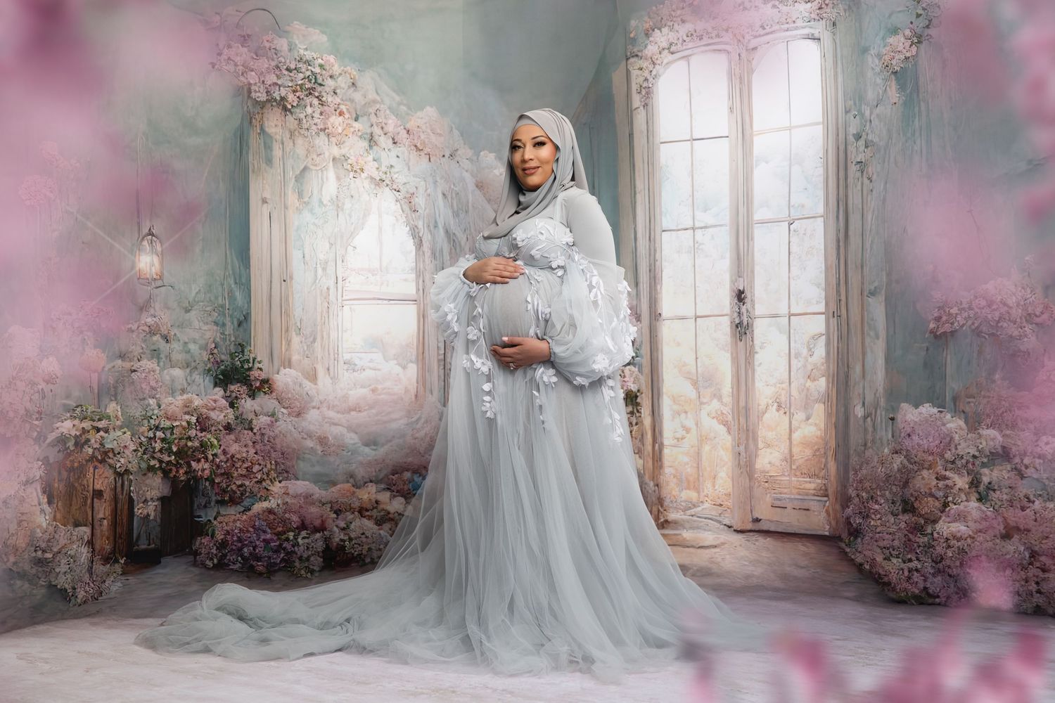 A pregnant woman in a flowing gray tulle gown poses in a dreamy floral setting with pink blossoms and French doors.