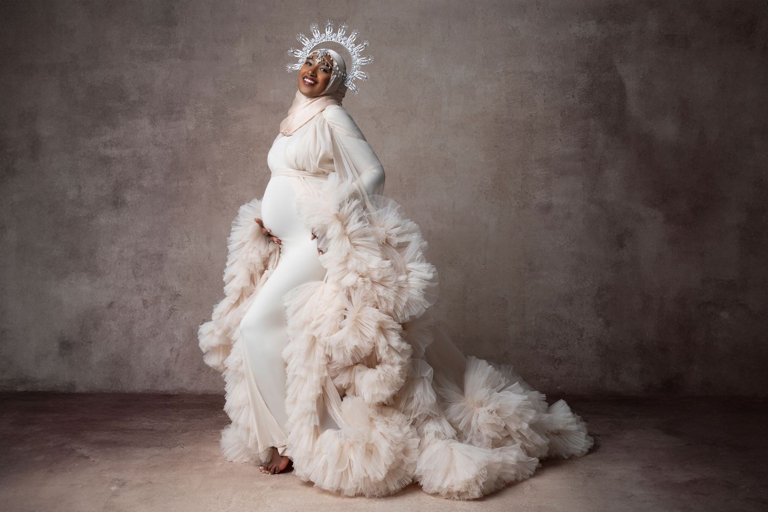 A person poses in an elegant white Victorian-style gown with voluminous ruffles and a dramatic flowing train against a gray backdrop.