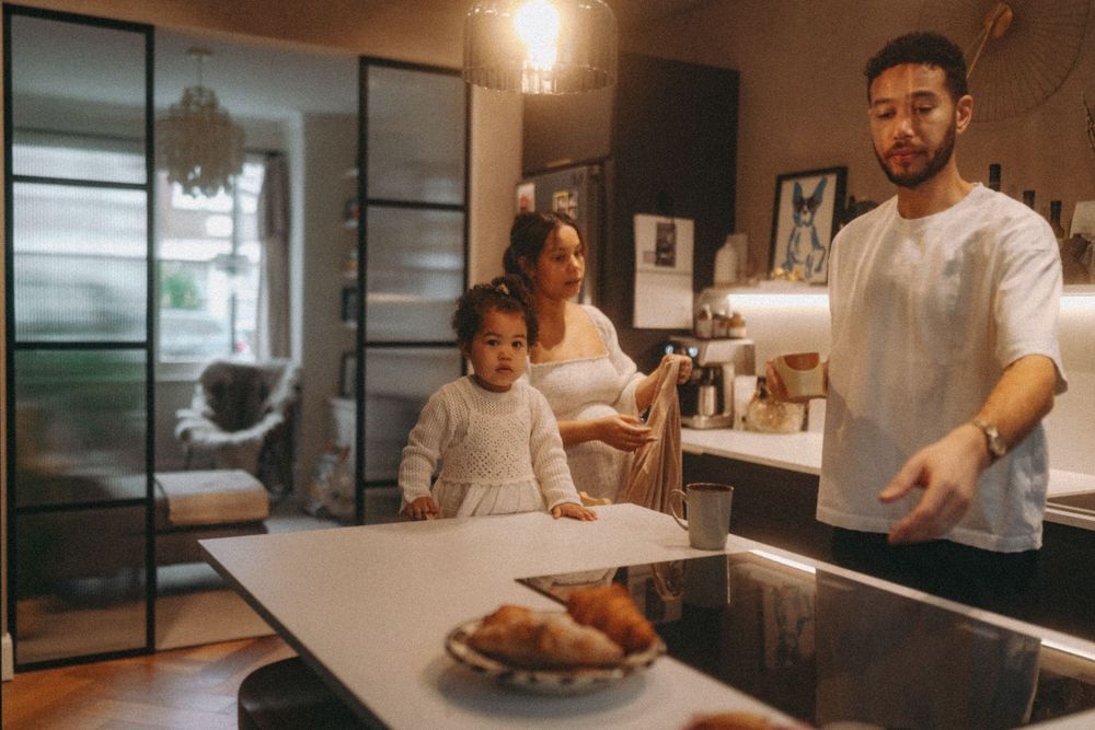 Candid moment between family of mum, dad & daughter in a kitchen with toddler looking towards camera. Family Photography