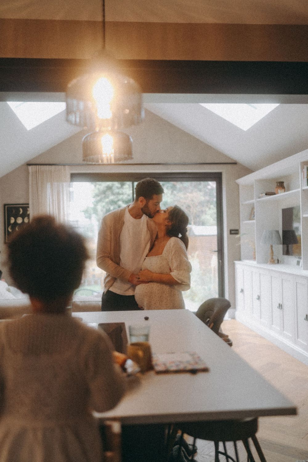 Couple sharing a kiss in a kitchen as toddler daughter looks on. Family documentary photography