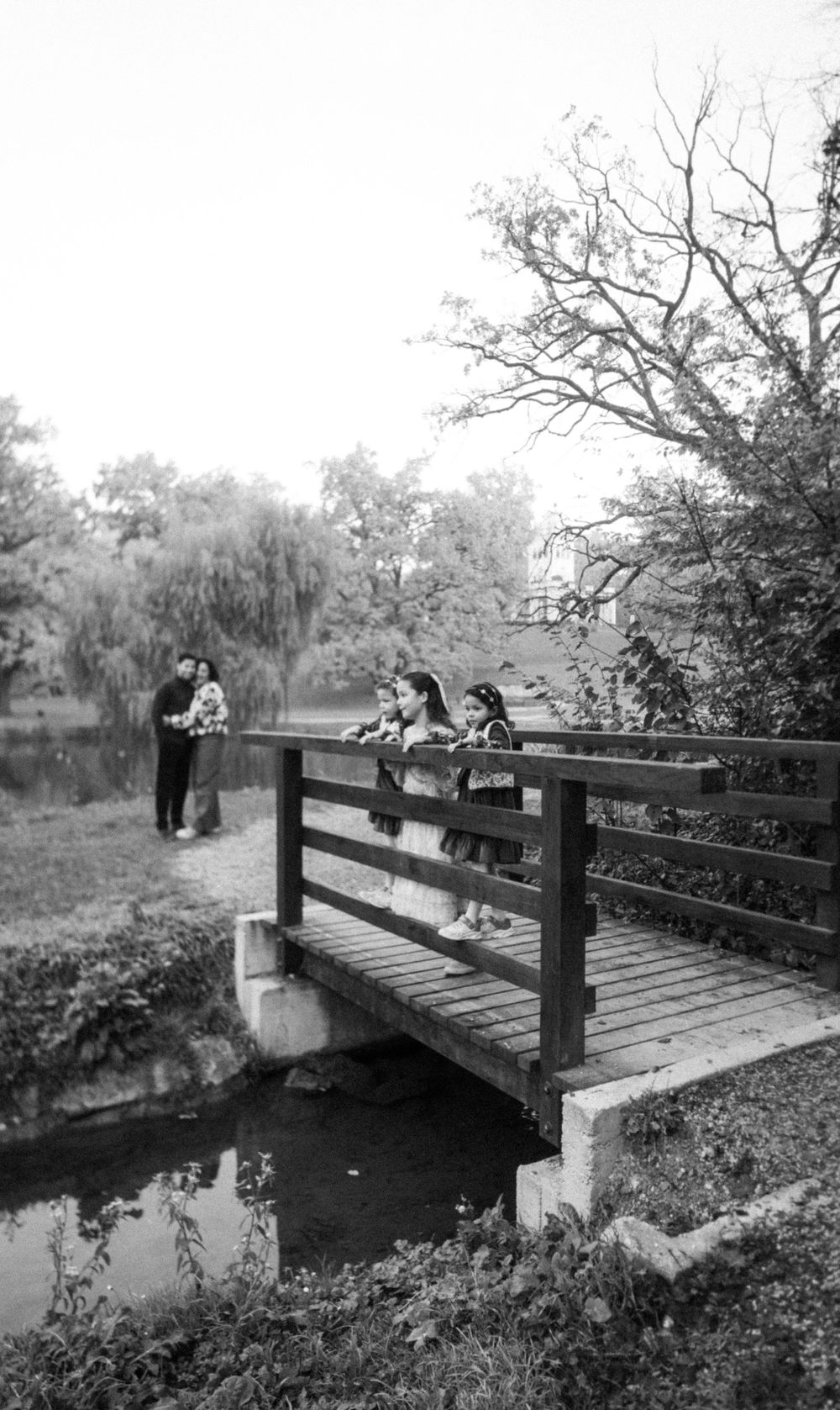 3 girls standing on a wooden bridge in a park looking at the stream as parents in an embrace far off look on. Family documentary photography