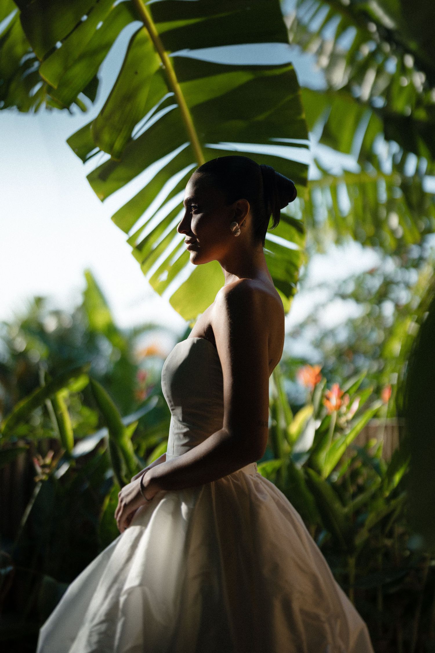 A silhouette stands in a white tulle skirt against tropical palm fronds in dramatic backlit natural lighting.