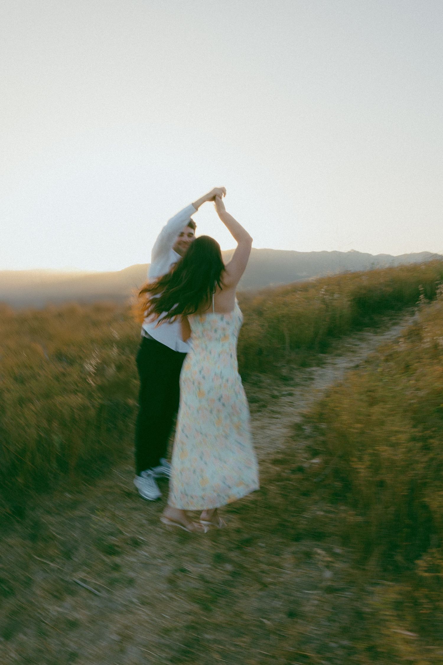 Two figures in light colored clothing dance together on a grassy hilltop against a sunset sky.