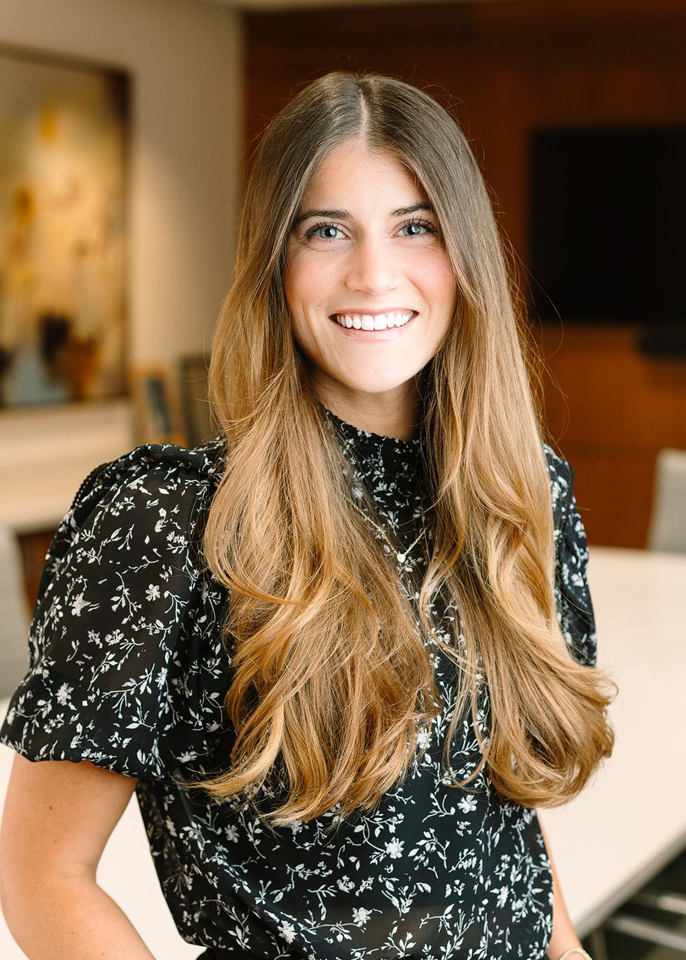 Elegant portrait of a person wearing a black and white floral print top with flowing honey-blonde hair and a bright smile.