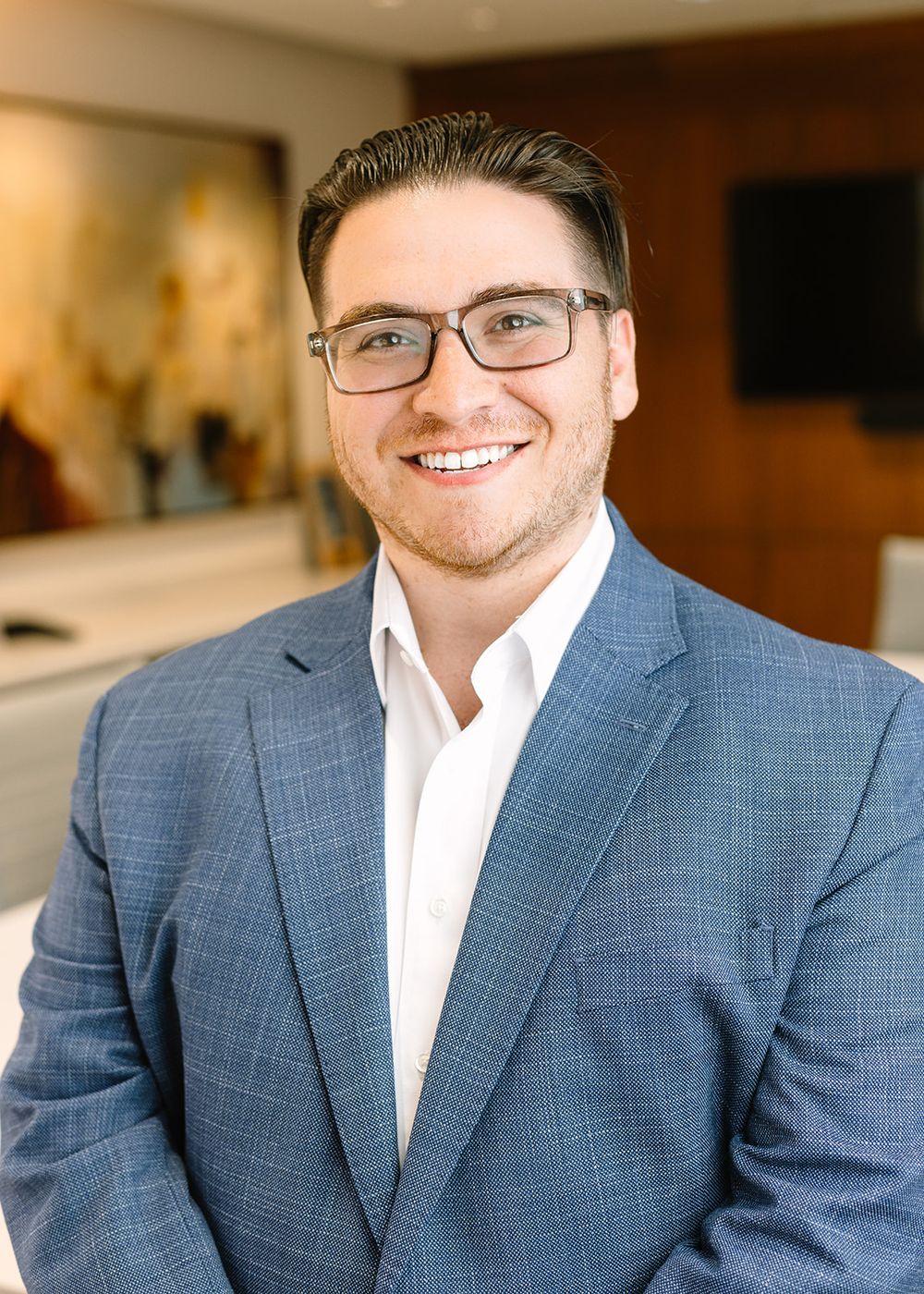 Professional wearing a blue-gray suit jacket and white dress shirt smiles warmly in an office setting.