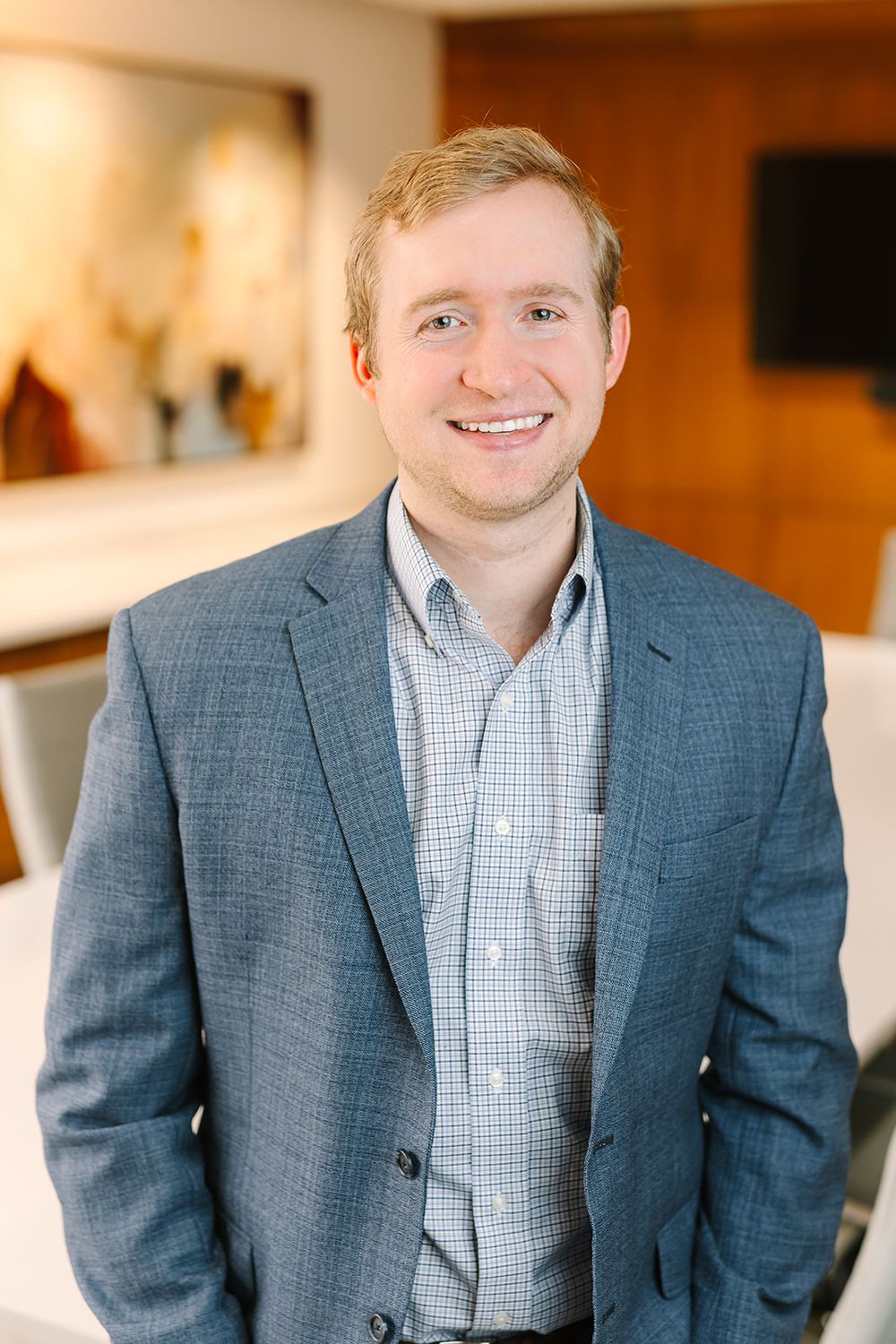 Professional wearing a blue suit jacket and checkered shirt smiles while posing in a warm interior setting.