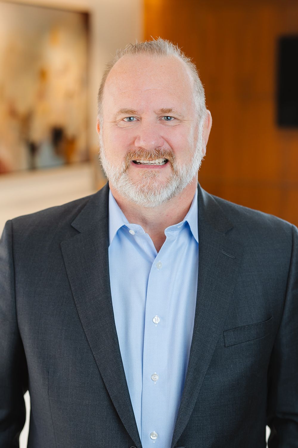Professional headshot of a bearded executive in a dark suit jacket and light blue button-down shirt against blurred office background.