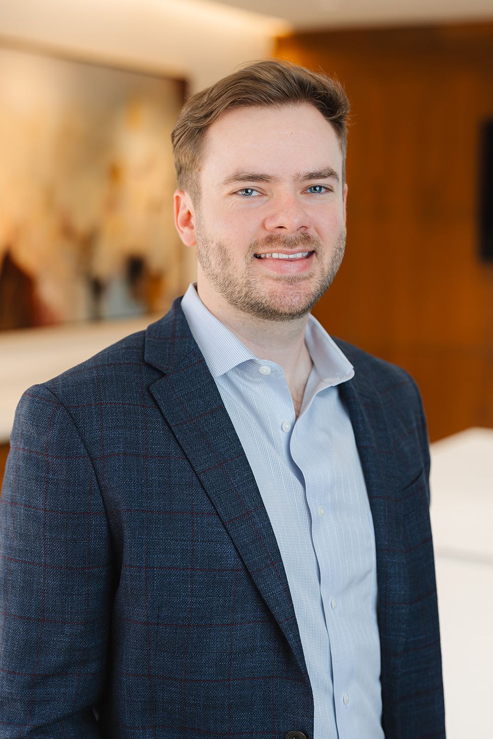 Professional headshot portrait with navy plaid suit jacket and light blue button down shirt in warm indoor setting.