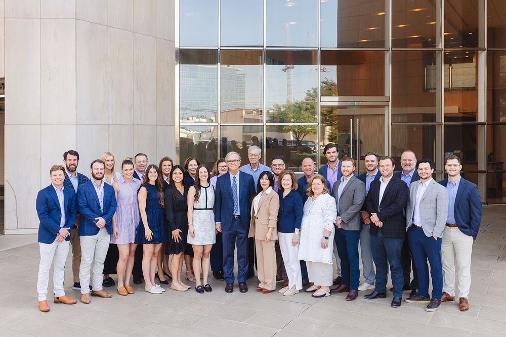 A professional group photo of office workers standing together outside a modern corporate building entrance.