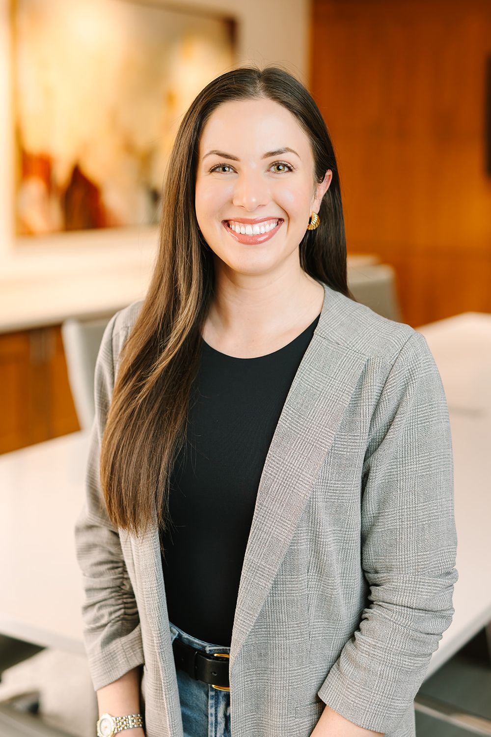 Professional headshot of someone in a gray blazer and black top smiling warmly in an office setting.