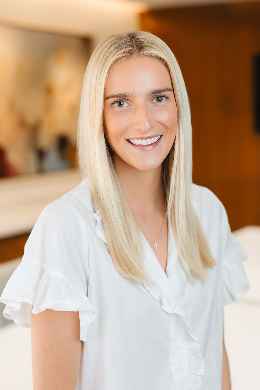 Portrait of someone with bright blue eyes and long platinum blonde hair wearing a white ruffled blouse in warm lighting.