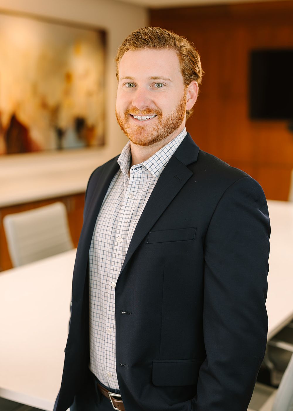 Professional wearing a navy suit jacket with checkered dress shirt and smiling warmly in corporate setting.