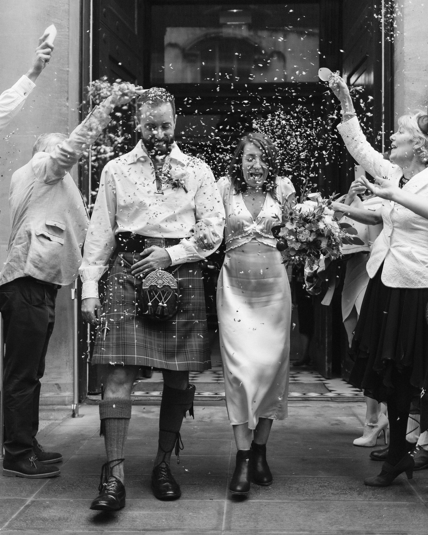 A wedding couple exiting a building while guests throw confetti in a joyful black and white celebration scene.