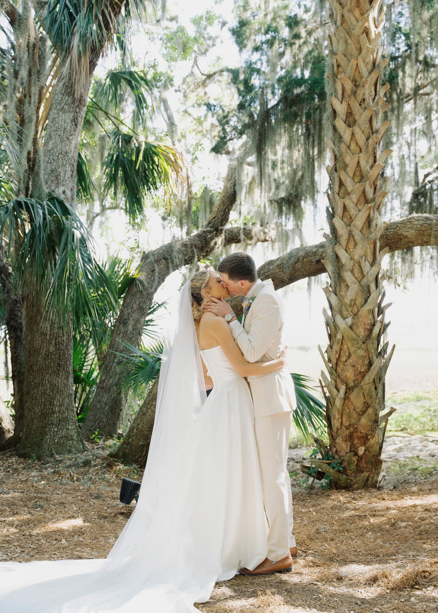 Bride and groom sharing first kiss under Spanish moss-draped trees in lush outdoor ceremony.