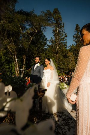 A romantic outdoor wedding ceremony takes place in a wooded setting during golden hour.