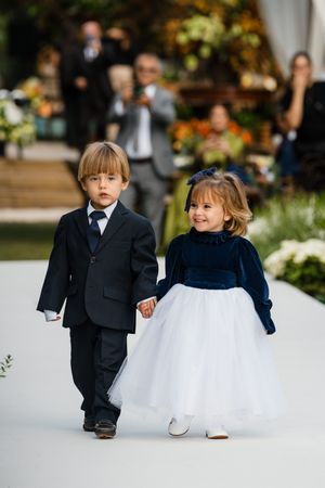 Two young children in formal attire walk hand in hand during a wedding ceremony on a white aisle.