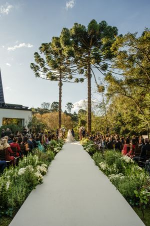 Outdoor wedding ceremony setup with white aisle runner between natural garden landscaping and tall Araucaria trees.