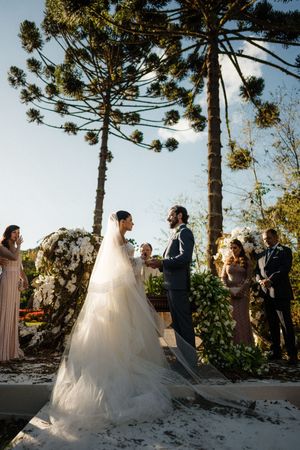 Wedding ceremony takes place outdoors with tall araucaria pine trees and sunlight creating a dreamy backdrop.