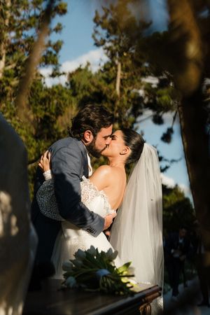 A romantic wedding couple shares an intimate kiss outdoors in front of pine trees at sunset.