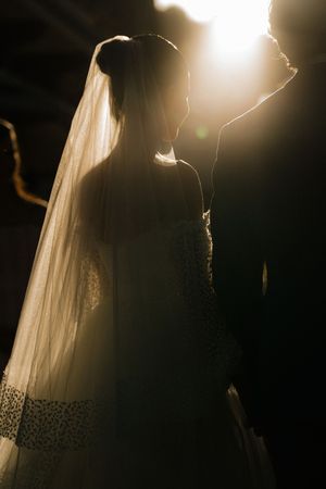 Silhouetted figures in elegant wedding attire bathed in dramatic backlit glow with flowing veil creating ethereal atmosphere.