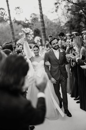 A newly married couple walk down an aisle lined with guests as the bride triumphantly holds up her bouquet in black and white.