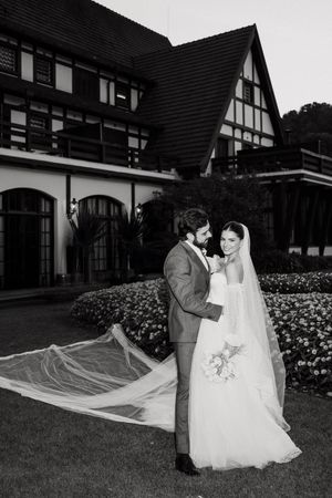 Newlyweds pose together on lawn in front of tudor-style building with bride wearing flowing veil and white gown.