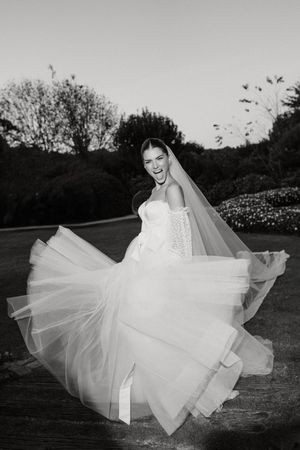 A bride in a flowing white tulle wedding dress twirls joyfully in a garden at dusk, captured in black and white.
