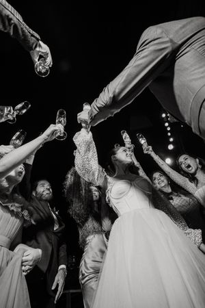 Black and white photo of wedding celebration with guests raising their glasses in a toast around someone in a white dress.