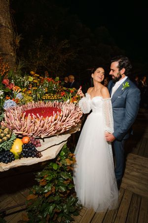 A couple in wedding attire share a moment next to a large floral display with fruits and protea flowers on a wooden deck.