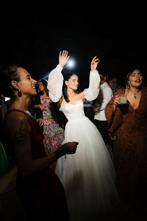 A bride in a white gown dances with her hands raised on the dance floor surrounded by celebrating wedding guests.