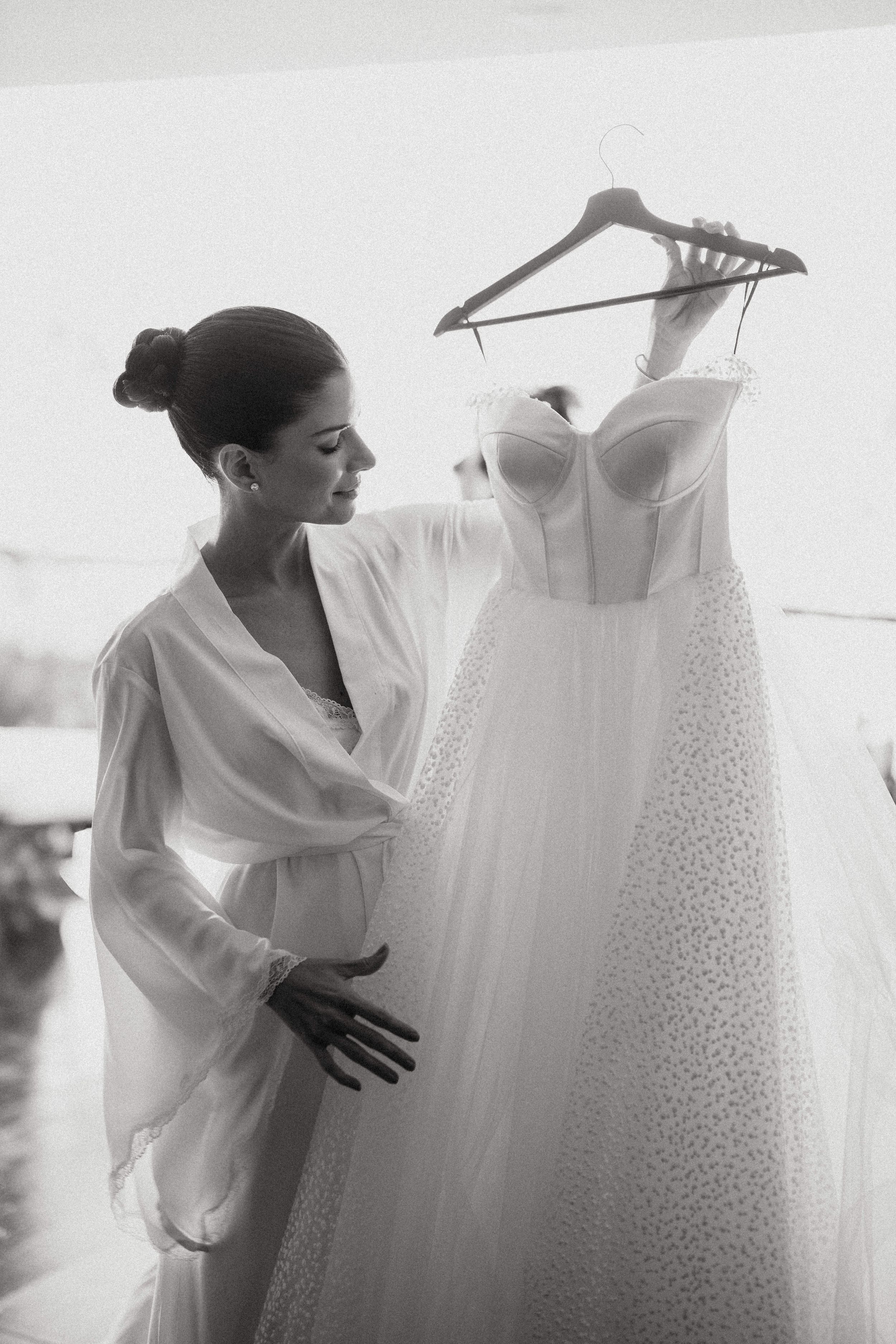 A bride in a white robe admires her elegant wedding dress with delicate details hanging on a hanger in black and white.