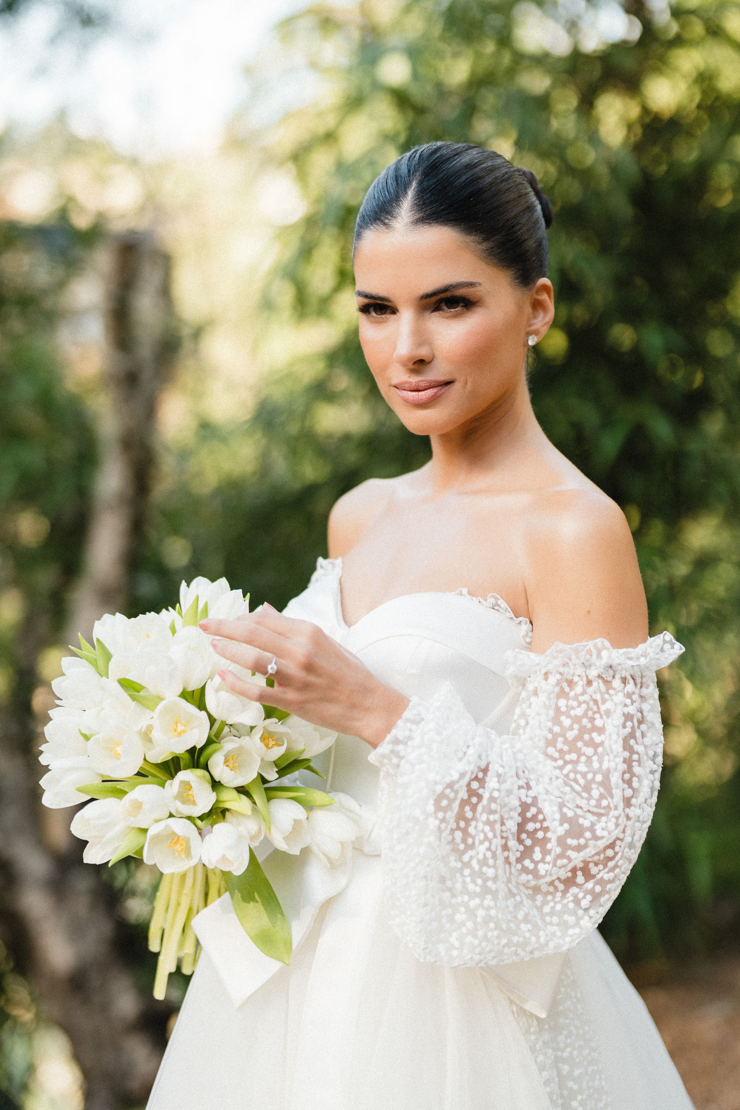 Bride in off-shoulder lace wedding dress holds elegant white tulip bouquet against blurred outdoor garden background.