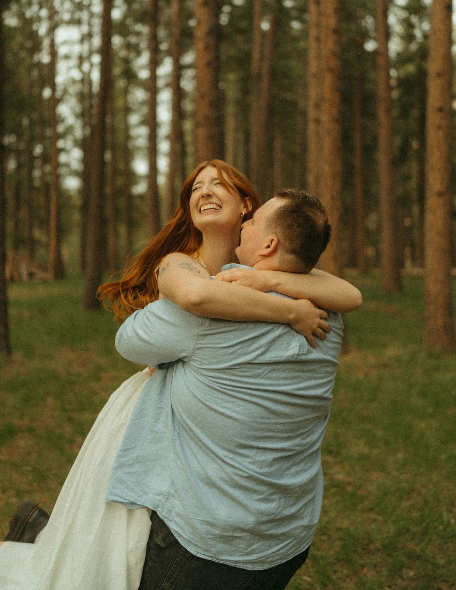 A couple shares a joyful embrace in a serene pine forest setting during golden hour.